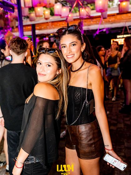 Dos amigas posan juntas en medio de la fiesta, con las luces cálidas y la gente detrás.