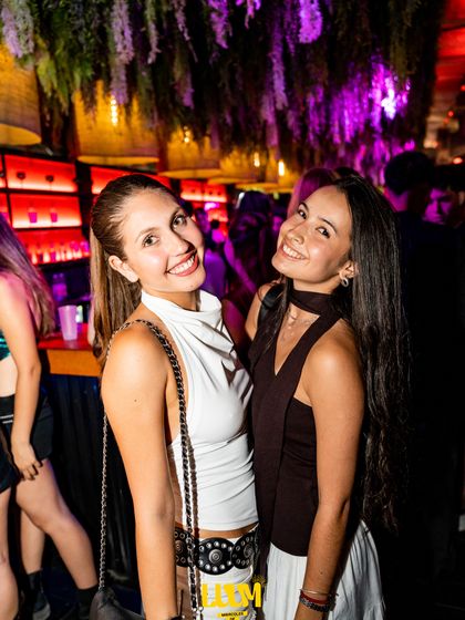 Dos amigas posan abrazadas entre luces moradas y cálidas.