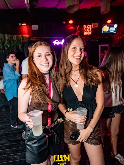 Dos amigas con sus vasos posan frente a un letrero de neón.