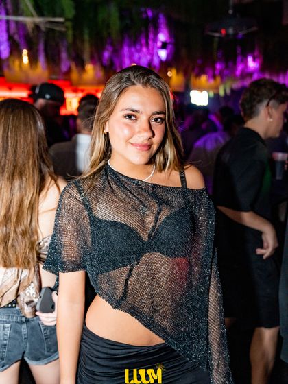 Una joven posa entre la gente, con las luces moradas del techo detrás.