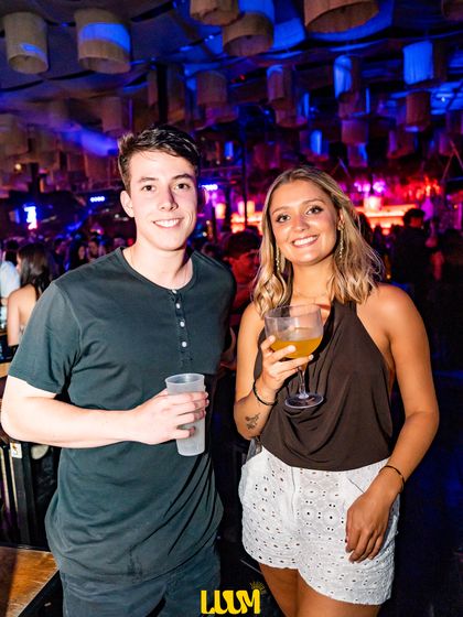 Una pareja con sus tragos posa bajo luces azules y moradas.