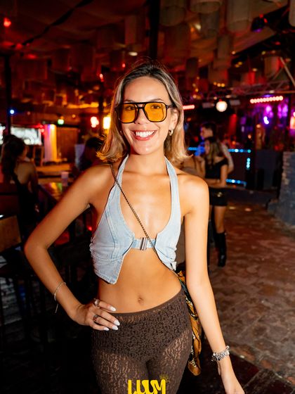 Una joven con lentes de sol posa con las manos en la cintura, rodeada de luces de colores.