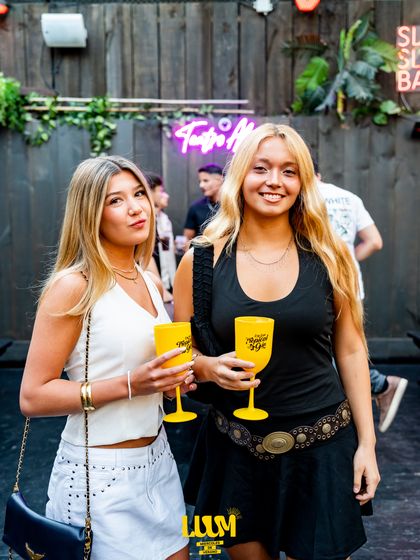 Dos amigas brindan con vasos amarillos frente a la cámara, en el sector de la barra.