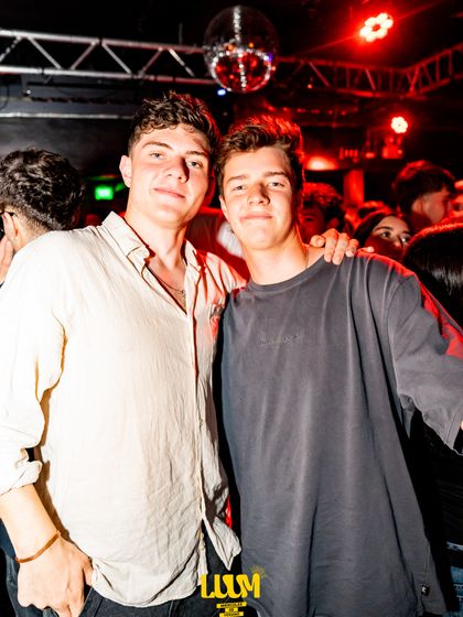 Dos amigos posan abrazados en medio del salón, con luz roja y gente alrededor.