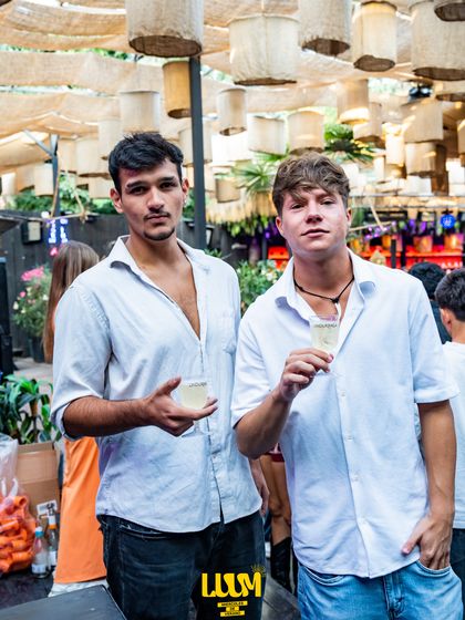 Dos amigos de camisa blanca brindan bajo las lámparas de mimbre del techo.