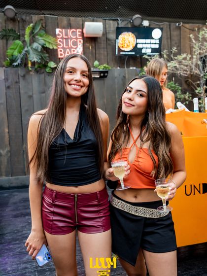 Dos amigas con copas de vino posan frente a un muro de madera con el letrero del local.