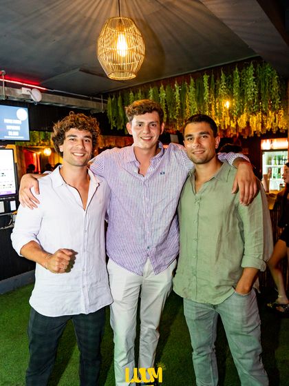 Tres amigos posan frente al muro verde del local, bajo una lámpara colgante.