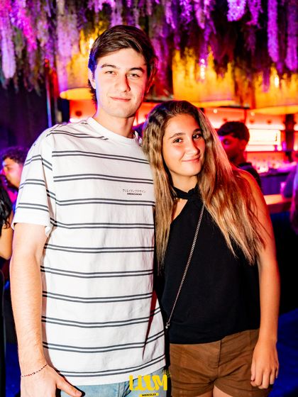 Dos jóvenes posan juntos bajo las plantas colgantes y las luces moradas del techo.