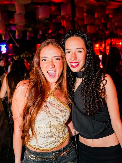 Dos amigas se ríen frente a la cámara, con la luz roja de la fiesta detrás.