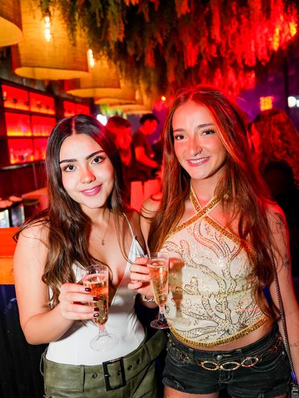 Dos amigas brindan con copas de champaña, con la barra iluminada de rojo al fondo.