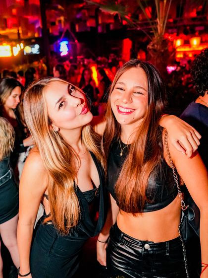 Dos amigas se abrazan riéndose, entre luces rojas y moradas.