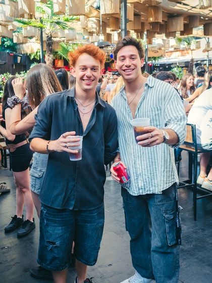 Dos amigos con sus tragos posan en la terraza al aire libre, con la gente y las mesas detrás.