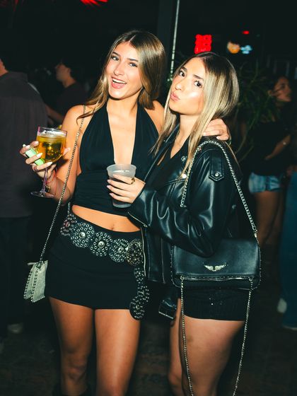Dos amigas posan con sus tragos, una de ellas con chaqueta de cuero, en medio de la fiesta.